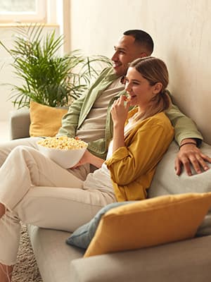 Young couple sat down eating popcorn in the front of the TV in their new home